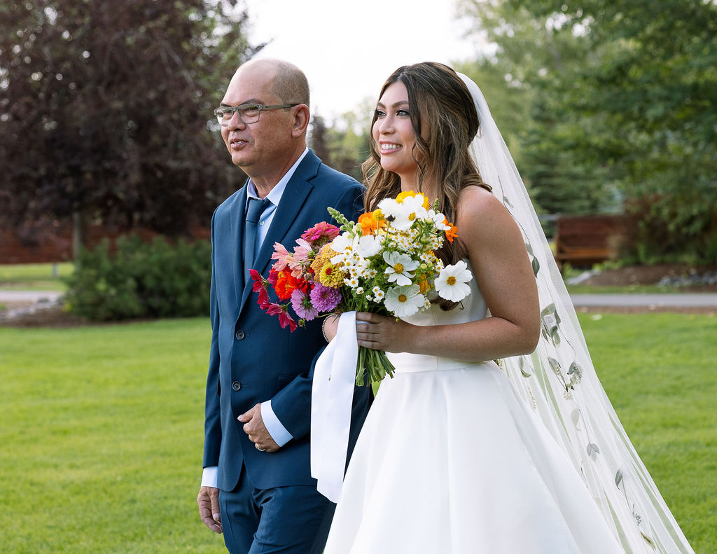 Bride and her father walk together through the garden lawn at Rockin’ TJ Ranch, the bride carrying a bright and colorful bouquet.