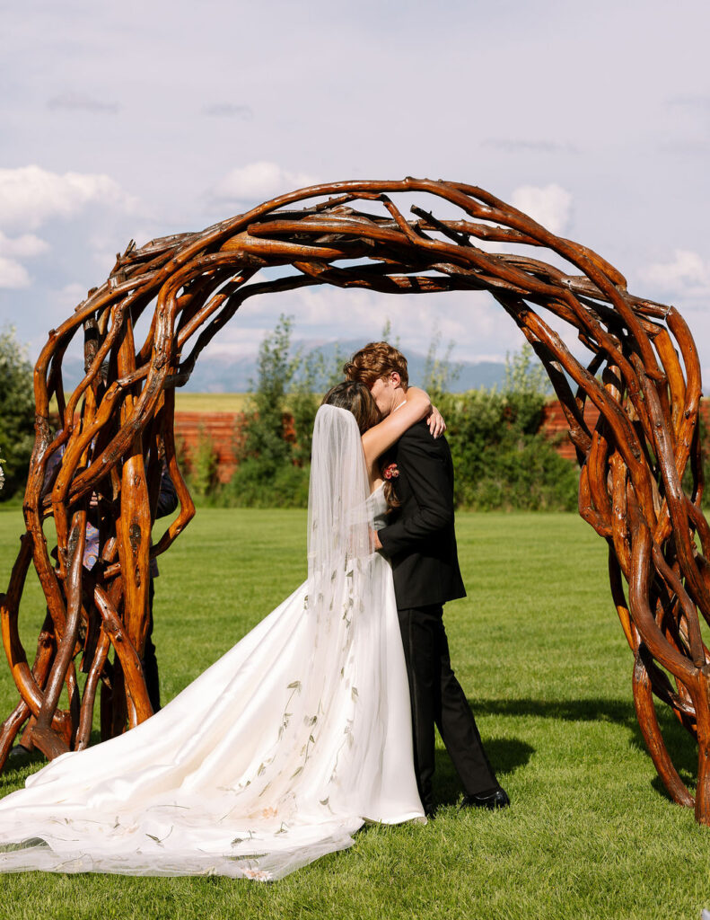 Bride and groom kiss beneath a rustic wooden ceremony arch with Montana mountain views at Rockin’ TJ Ranch in Bozeman.