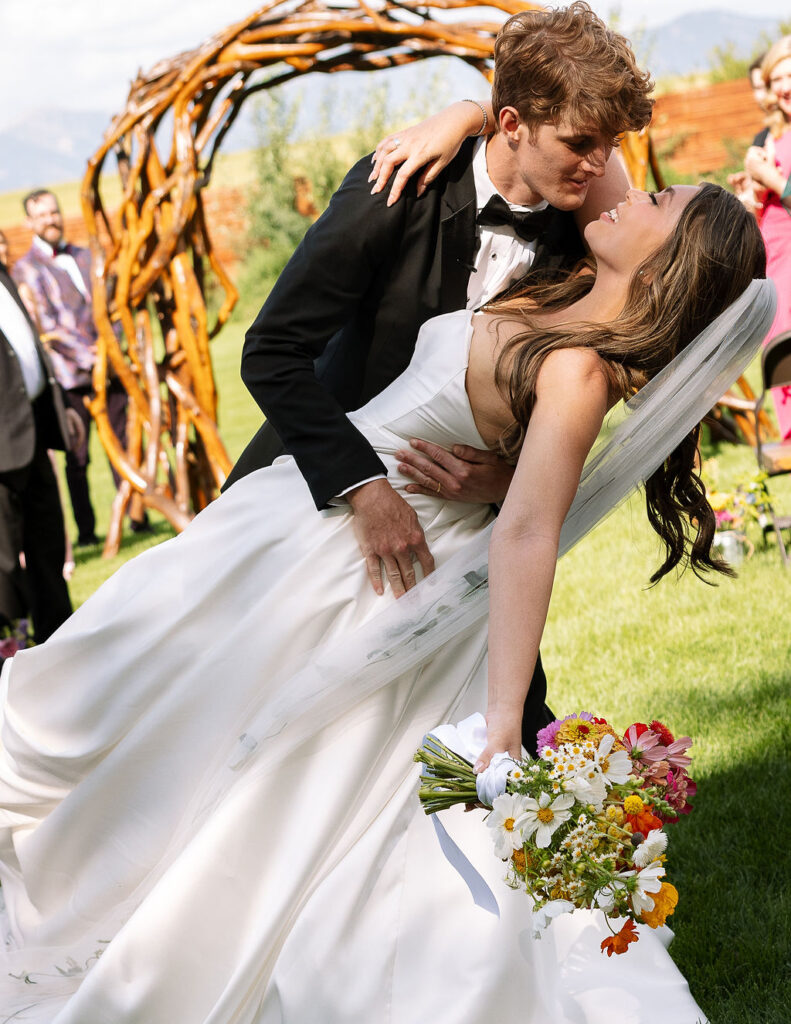 Bride and groom share a romantic dip and kiss on the lawn of Rockin’ TJ Ranch, her colorful bouquet adding a pop of color.
