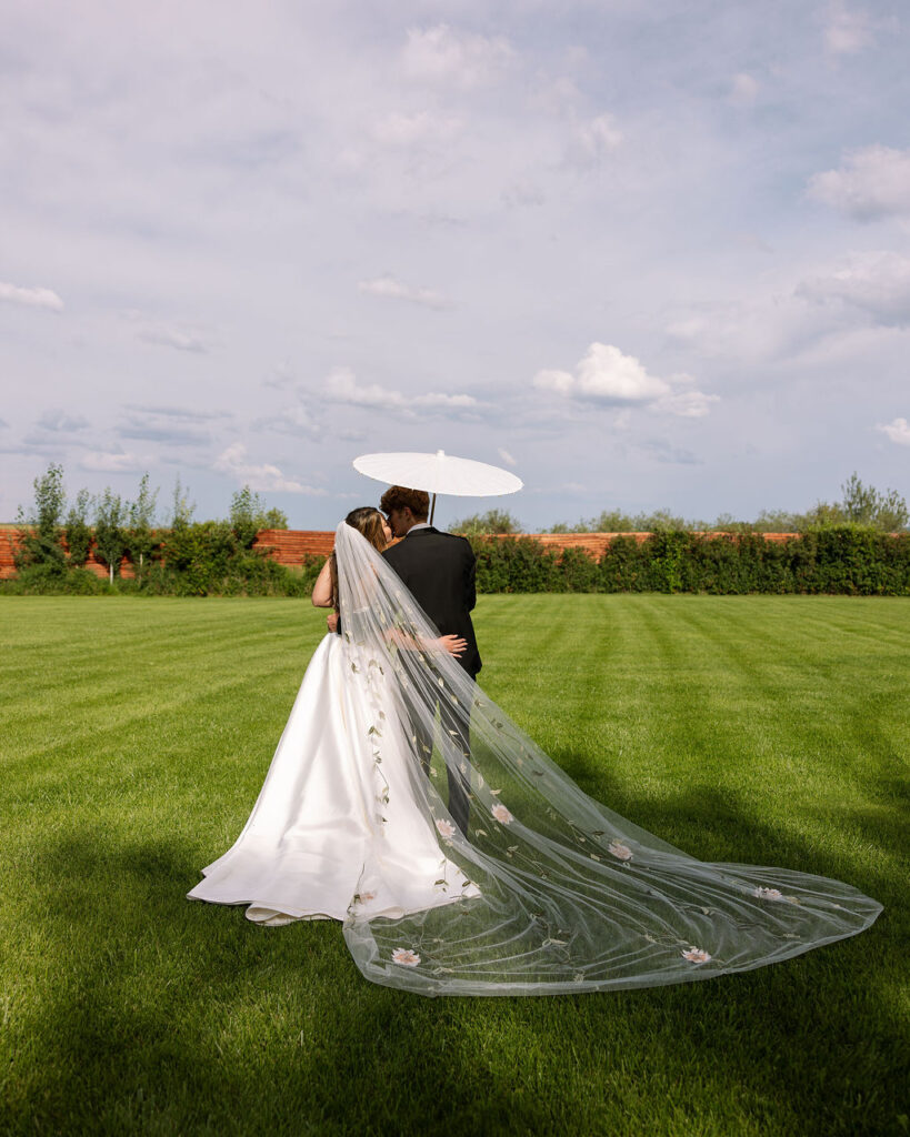 Bride walking with a flowing veil and long train while holding her bouquet, groom nearby with parasol at Rockin’ TJ Ranch in Bozeman.