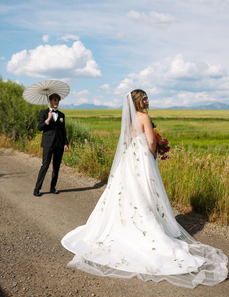 Groom holds a parasol over the bride as they share a kiss on a country road at Rockin’ TJ Ranch with the Bridger Mountains behind them.