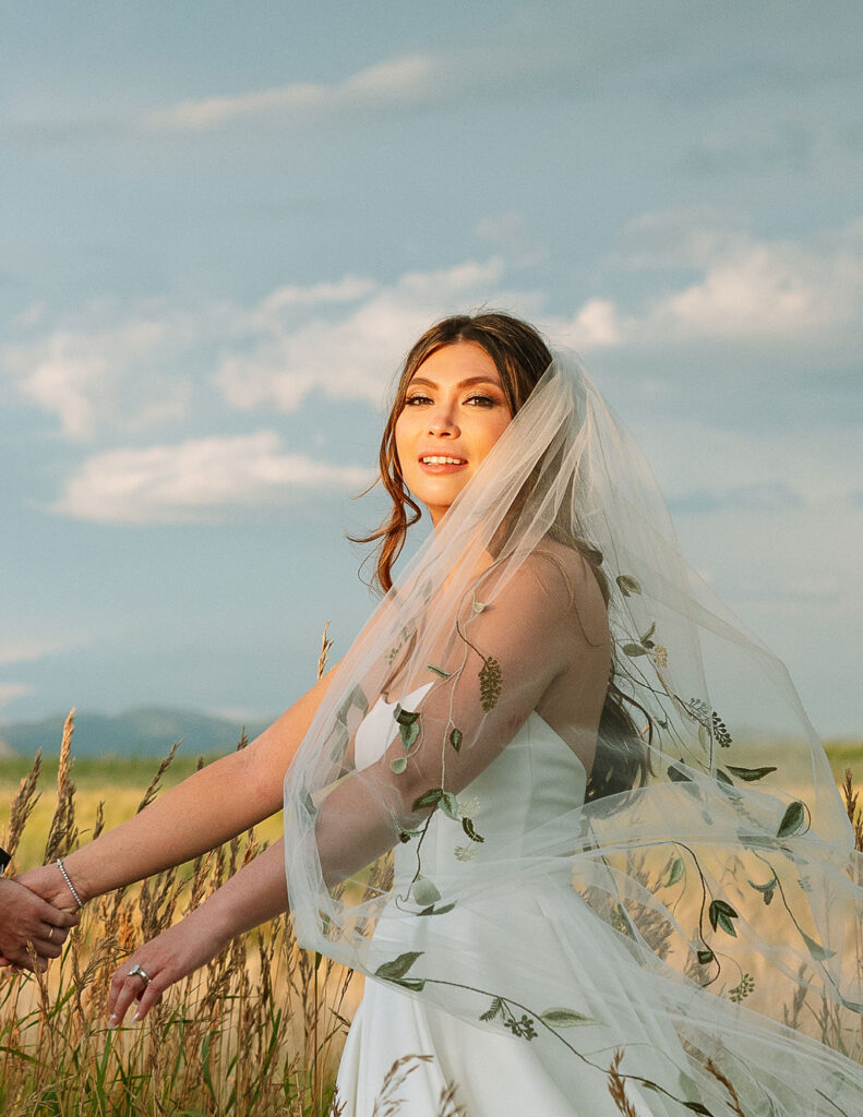 Bride smiling in her wedding dress with veil draped over her shoulders, surrounded by the golden fields of Rockin’ TJ Ranch in Bozeman.