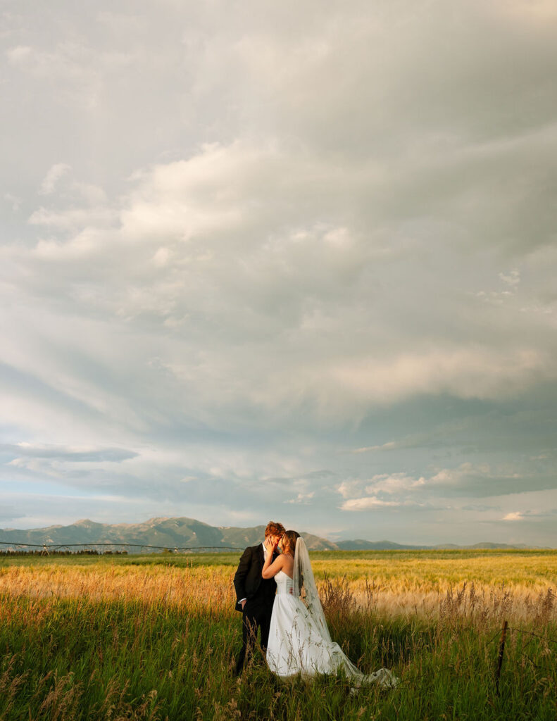 Couple standing in a wide-open Montana field at Rockin’ TJ Ranch with dramatic skies and the Bridger Mountains behind them.