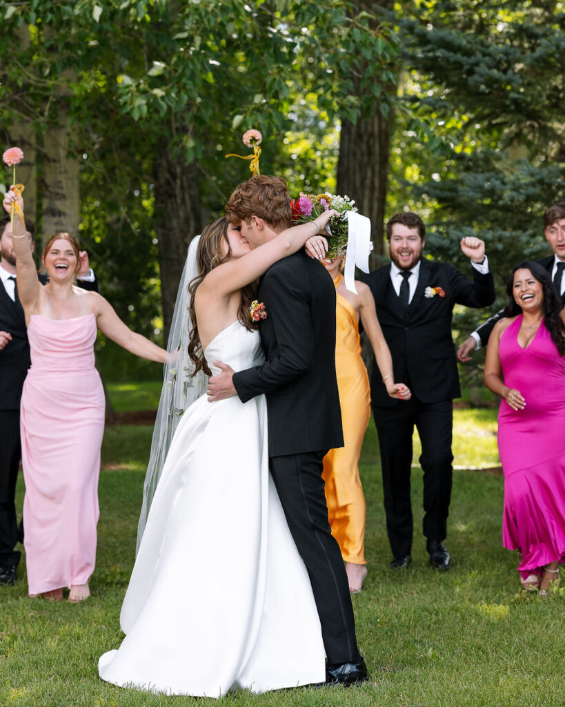Bride and groom share a celebratory kiss while wedding party cheers behind them on the lawn at Rockin’ TJ Ranch in Bozeman.