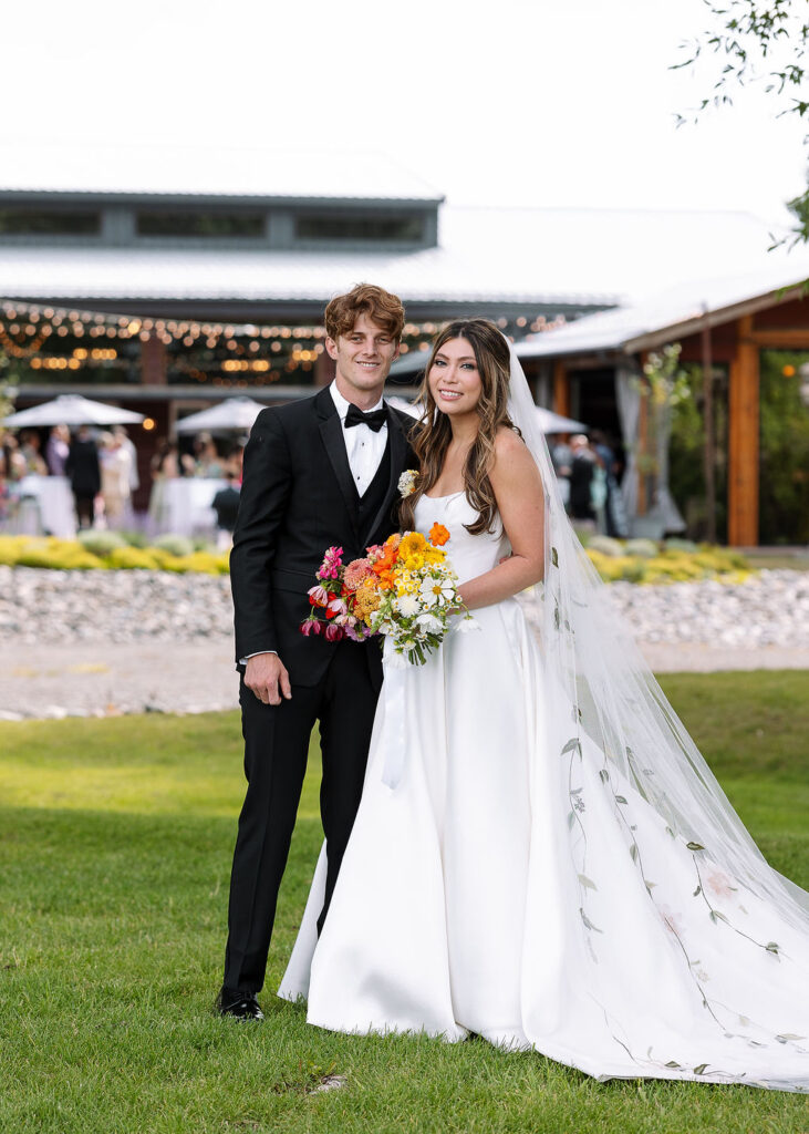 Bride and groom standing together outside Rockin’ TJ Ranch in Bozeman, holding a colorful bouquet and smiling.