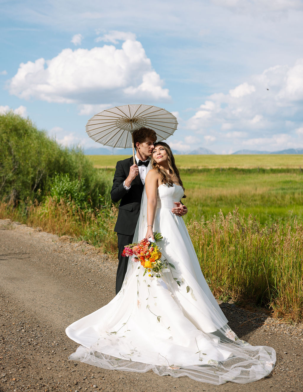 Bride and groom at their Rockin’ TJ Ranch wedding in Bozeman, Montana, sharing a romantic moment under a parasol with the Bridger Mountains in the background and the bride holding a colorful bouquet.