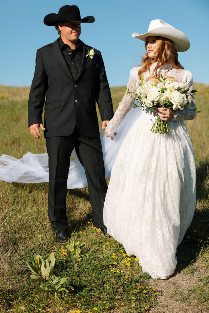 Bride and groom walking hand in hand through the grass at Clydesdale Outpost, her lace gown flowing behind as they smile at each other.