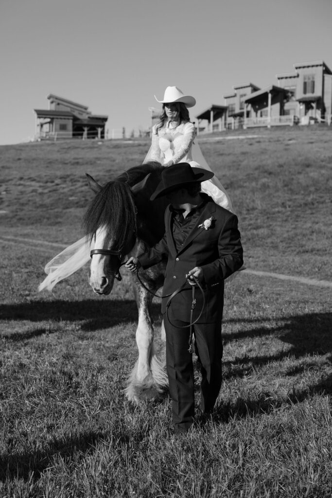 Bride riding a horse with her groom walking beside her across the rolling hills of Clydesdale Outpost during their Montana elopement.