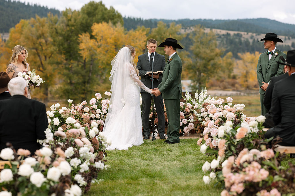 Bride and groom holding hands at floral ceremony backdrop in Montana