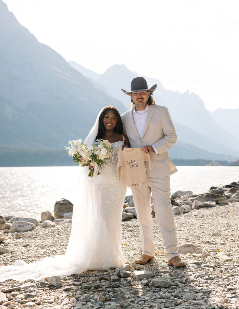 Couple standing along rocky lakeshore with mountains behind them during fall elopement in Glacier National Park