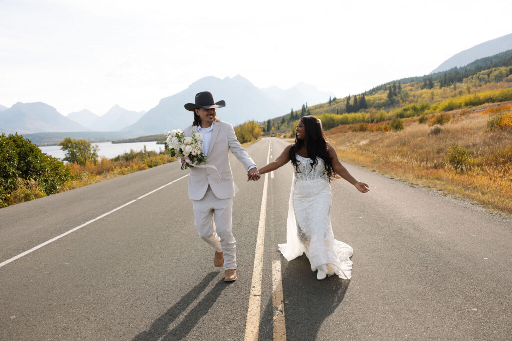 Couple running hand in hand down a scenic road in Glacier National Park during fall elopement