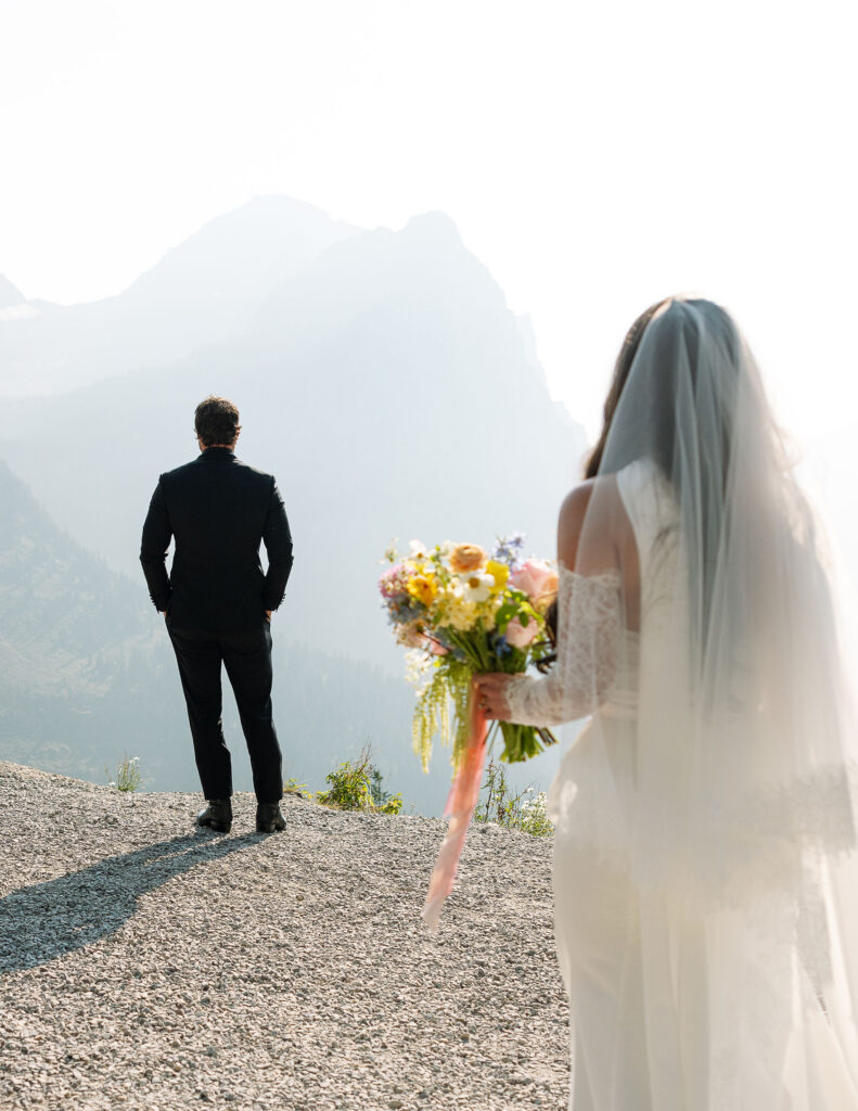 Bride walking toward groom holding a colorful bouquet with mountain views during a Glacier National Park elopement
