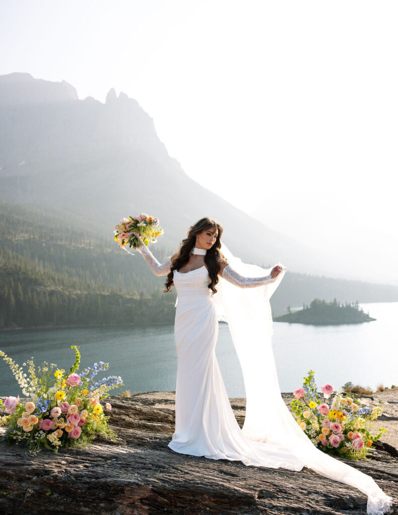 Bride standing on a cliffside overlooking a glacial lake with mountains behind her during a Glacier National Park elopement
