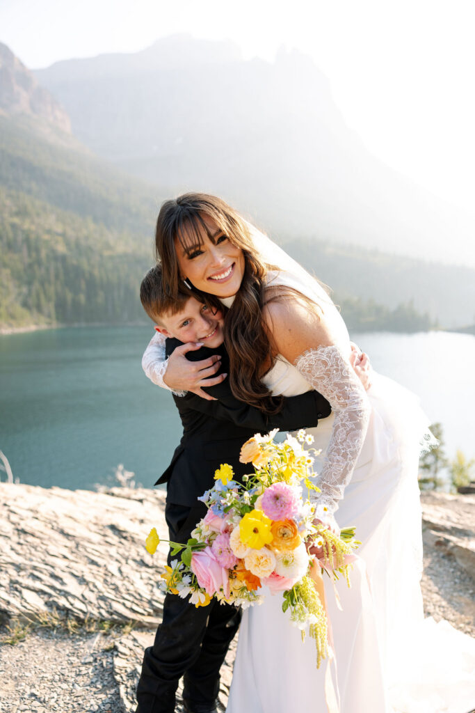 Bride hugging groom while holding a colorful bouquet beside a glacial lake in Glacier National Park
