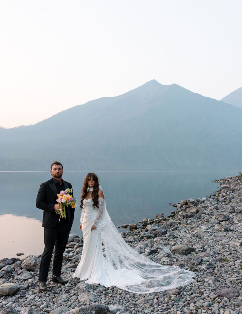 Bride and groom standing on a rocky lakeshore with mountain views during their Glacier National Park elopement