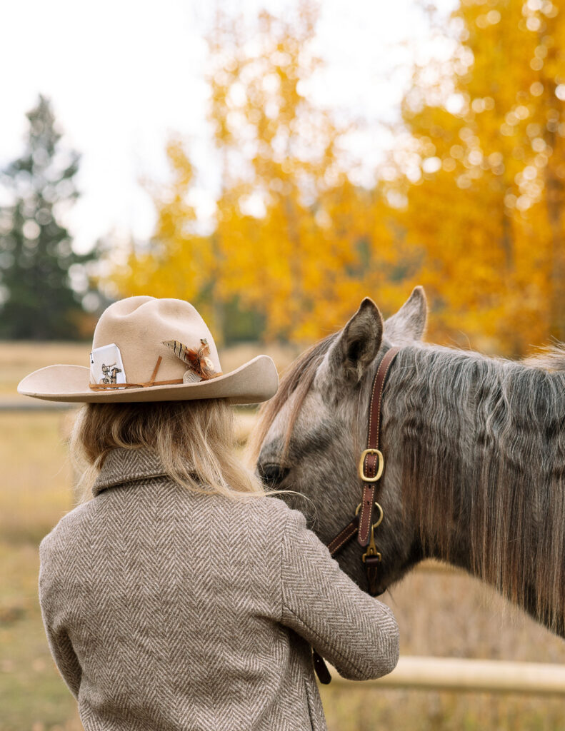 Haley Jessat in a cowboy hat standing beside her horse in a Montana field with golden fall foliage during a western photoshoot