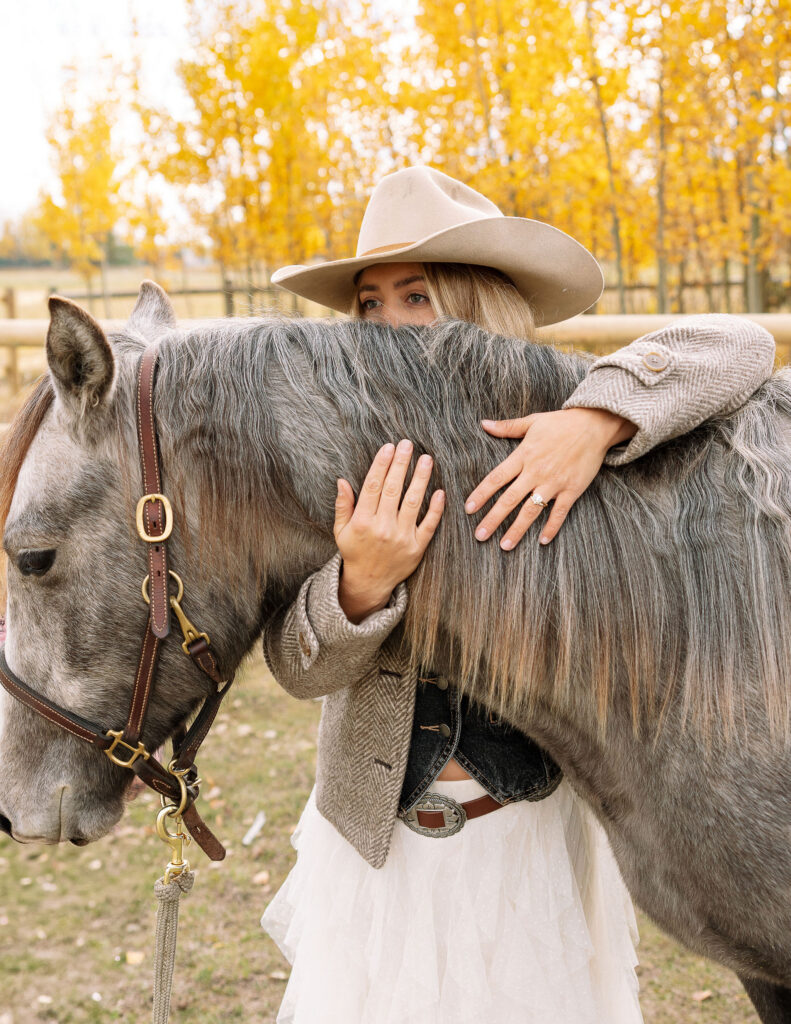 Haley Jessat in a beige western hat hugging a gray horse during a fall western photoshoot in Montana with golden autumn trees in the background