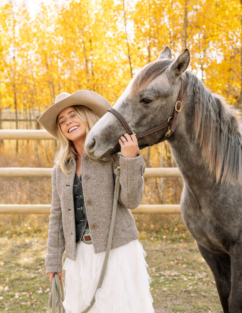 Haley Jessat standing beside a gray horse and smiling during a western photoshoot in Montana with autumn foliage and ranch fencing behind her