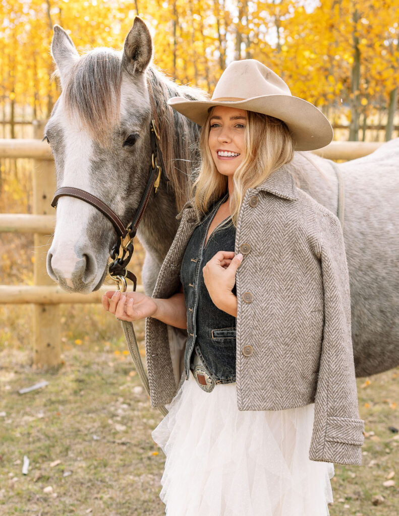 Haley Jessat smiling while holding the lead of her gray horse during a western portrait session in Montana with warm autumn colors