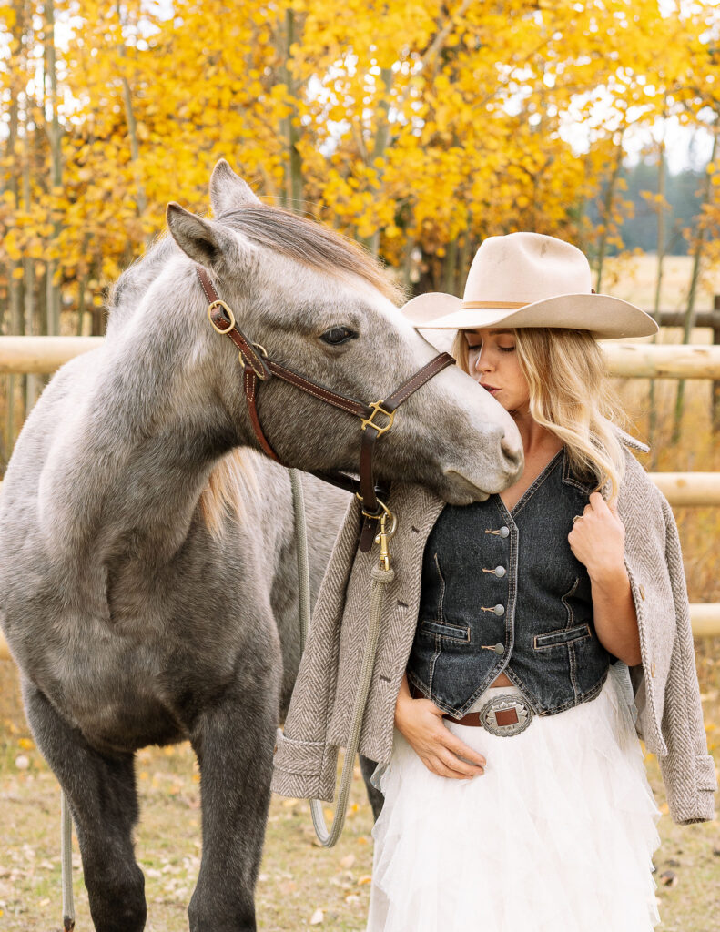 Gray horse nuzzling Haley Jessat during a western photoshoot in Montana with autumn foliage and rustic ranch fencing