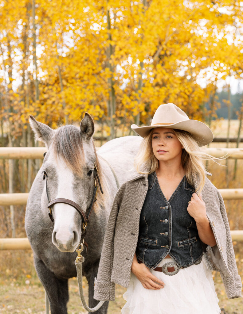 Haley Jessat standing beside a gray horse during a western lifestyle photoshoot in Montana with golden fall trees and ranch fencing in the background