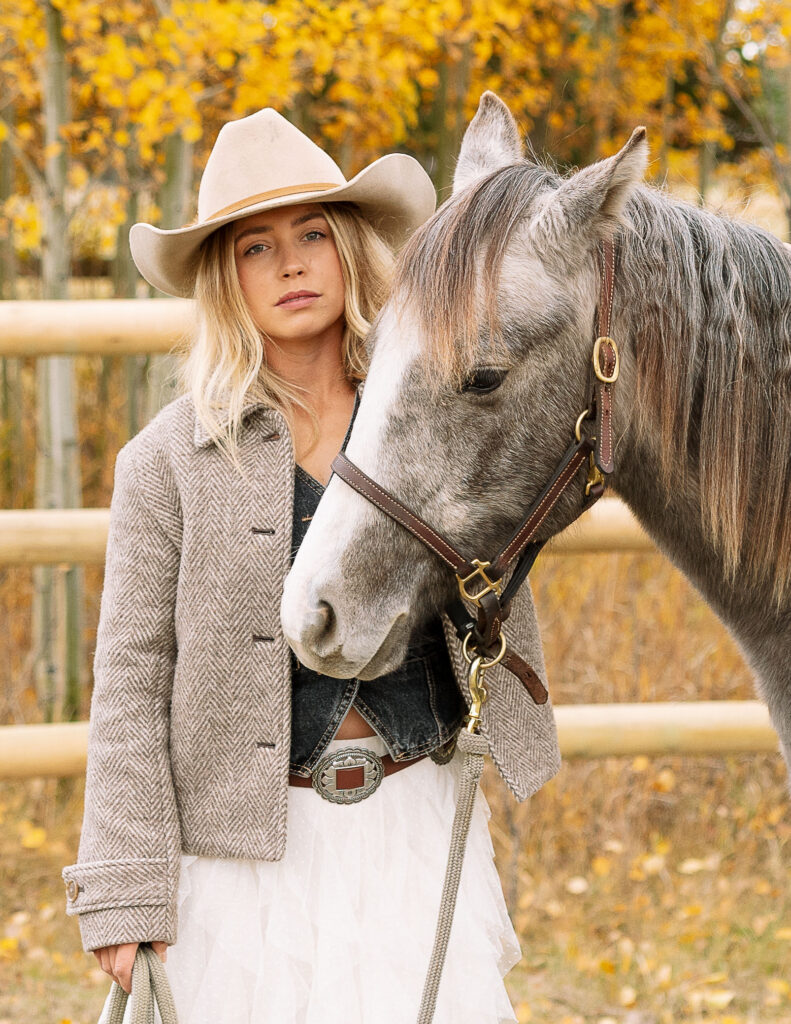 Haley Jessat standing beside her gray horse during a western lifestyle photoshoot in Montana with golden autumn trees
