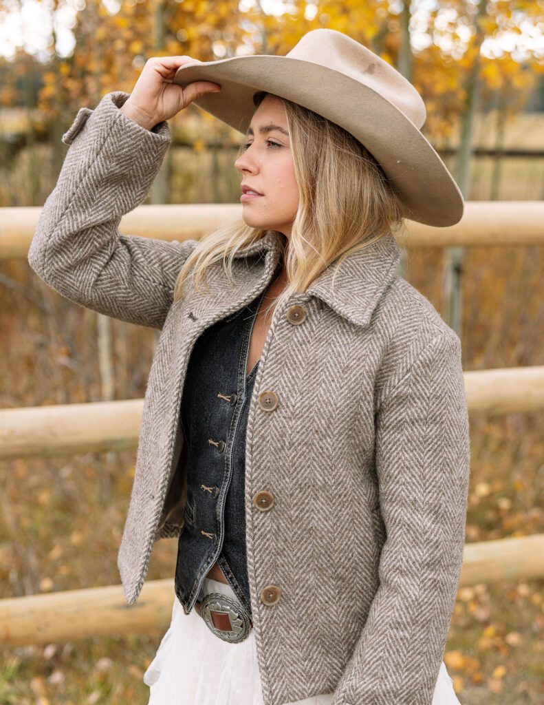 Haley Jessat wearing a western jacket and cowboy hat during a Montana ranch photoshoot with rustic fencing and autumn trees