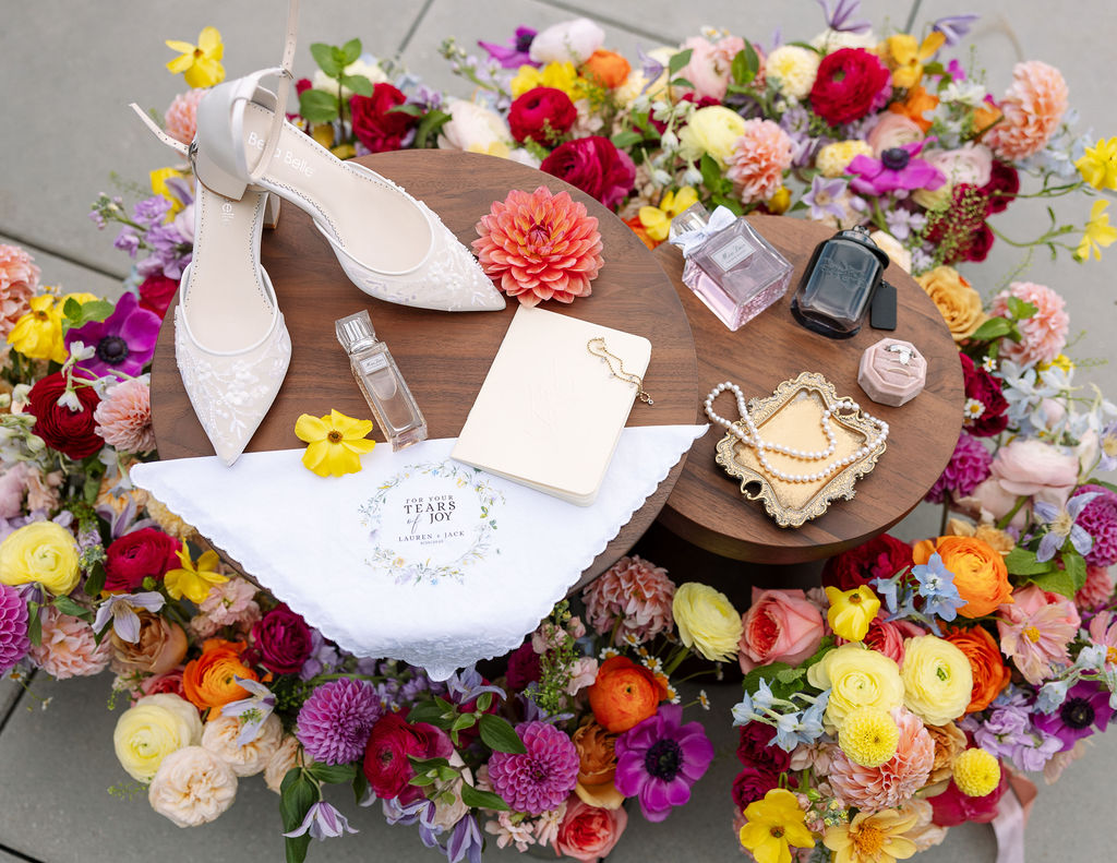 Wedding detail flat lay with bridal shoes, rings, perfume, and colorful florals arranged around the invitation suite
