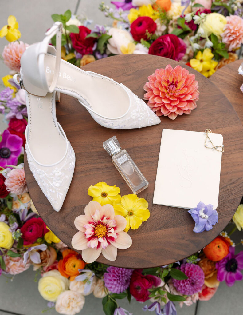 Wedding detail flat lay featuring bridal shoes, perfume, florals, and invitation suite
