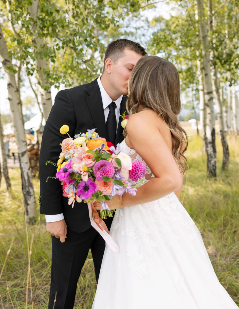 Bride and groom standing together in a grove of trees during sunset portraits while holding a colorful bouquet