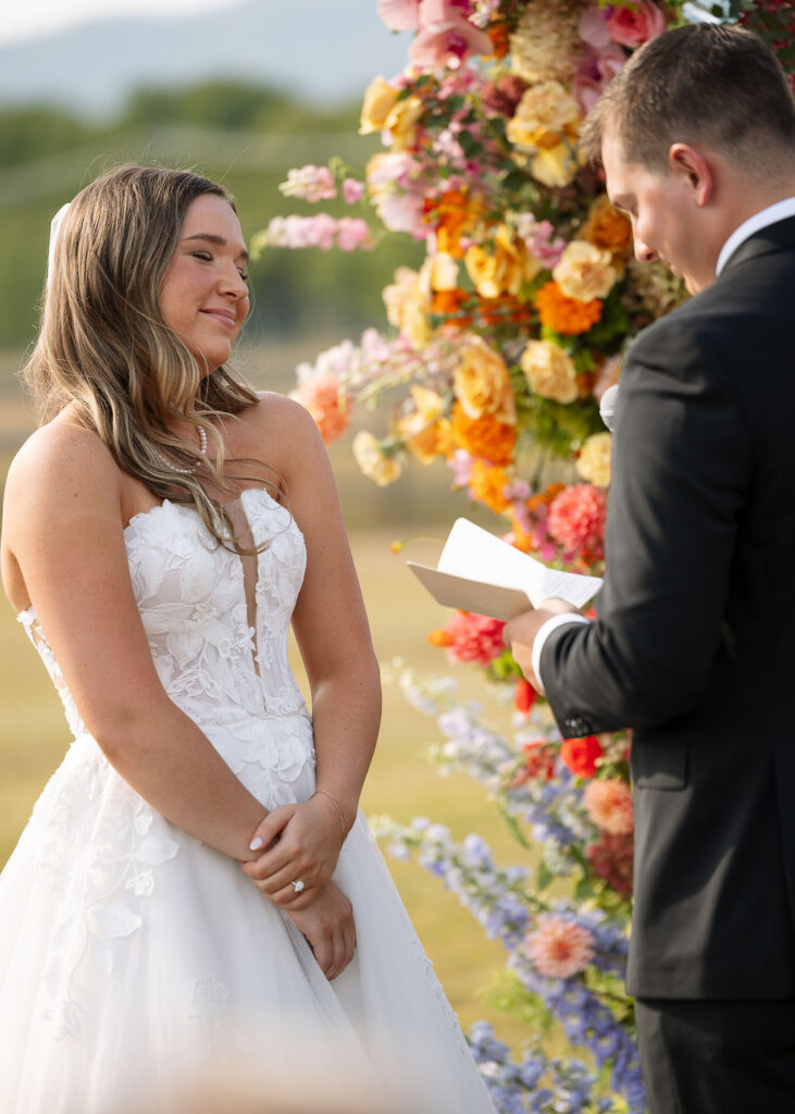 Bride smiling during an outdoor wedding ceremony while groom reads vows beneath colorful floral arrangements