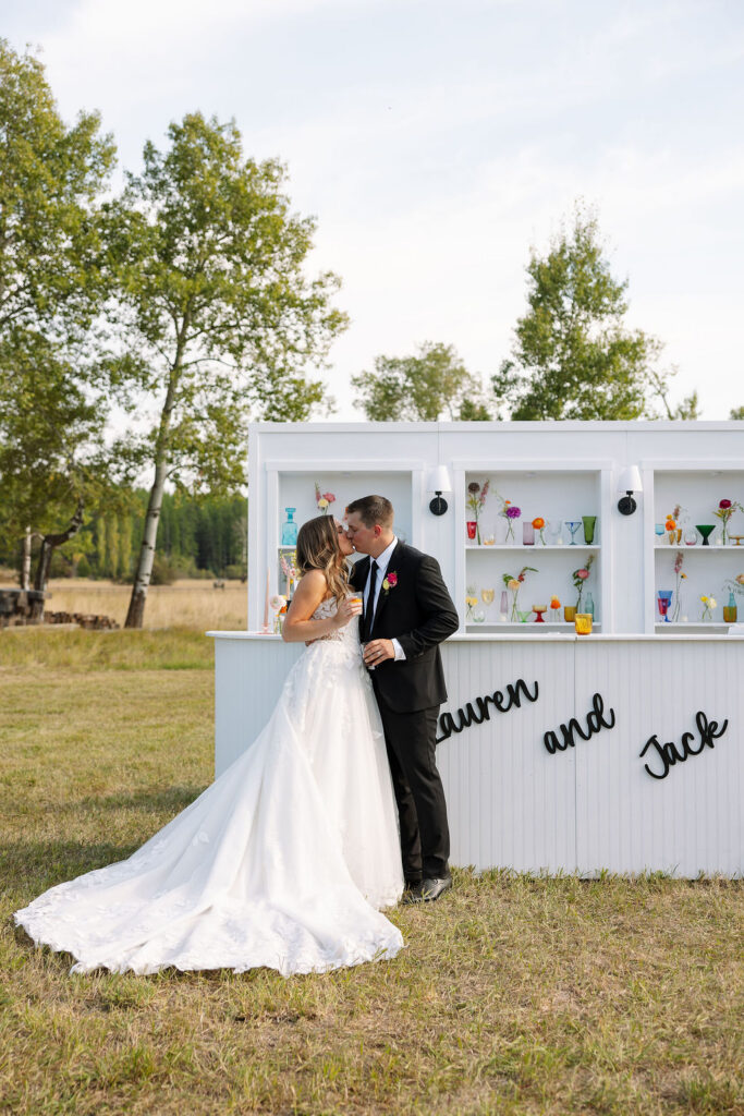 Bride and groom sharing a kiss beside a custom white bar during an outdoor summer wedding reception in Whitefish Montana