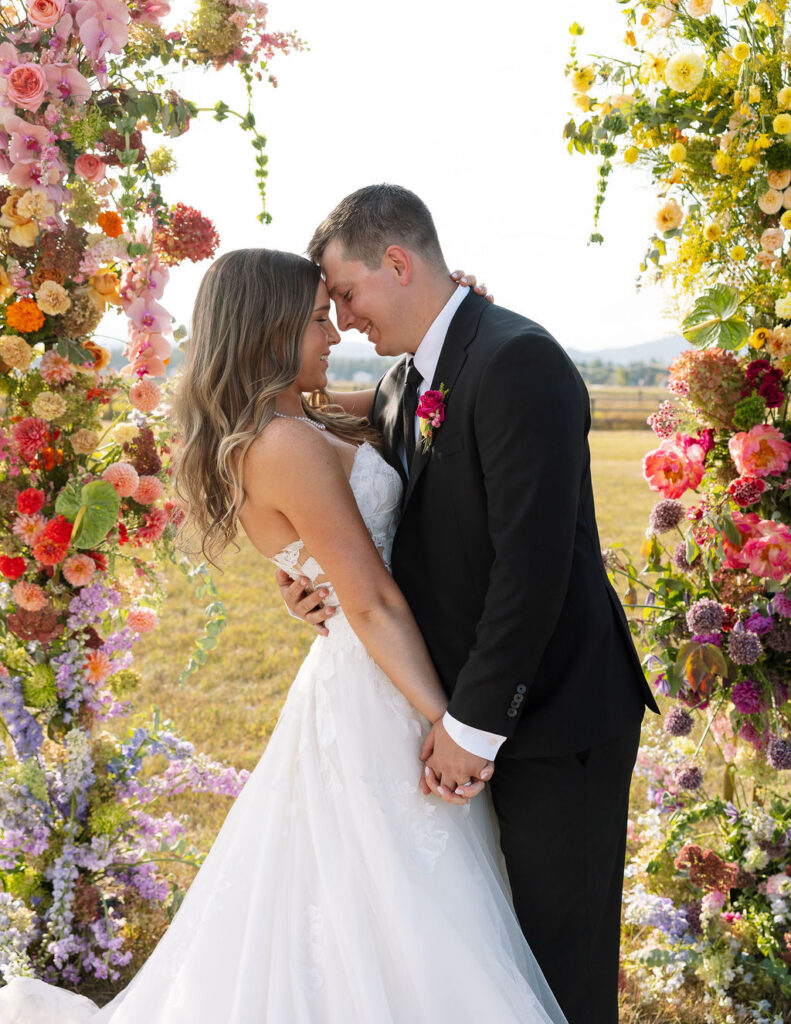 Bride and groom standing together beneath a colorful floral ceremony installation during sunset portraits in Whitefish Montana