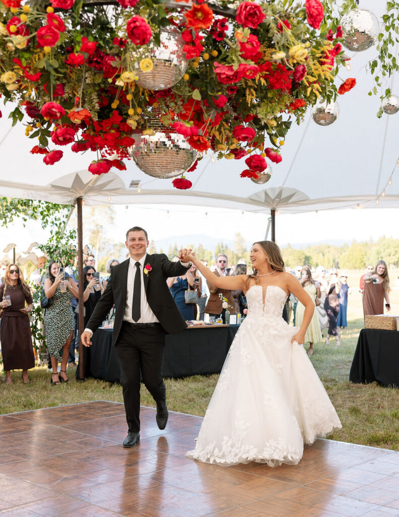 Bride and groom running onto the dance floor under a vibrant floral installation during their summer wedding reception in Whitefish Montana