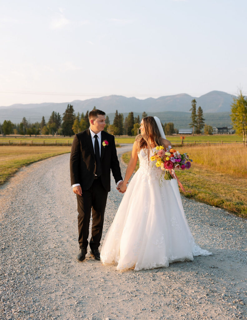 Bride and groom walking down a gravel road together during sunset portraits with mountain views in Whitefish Montana