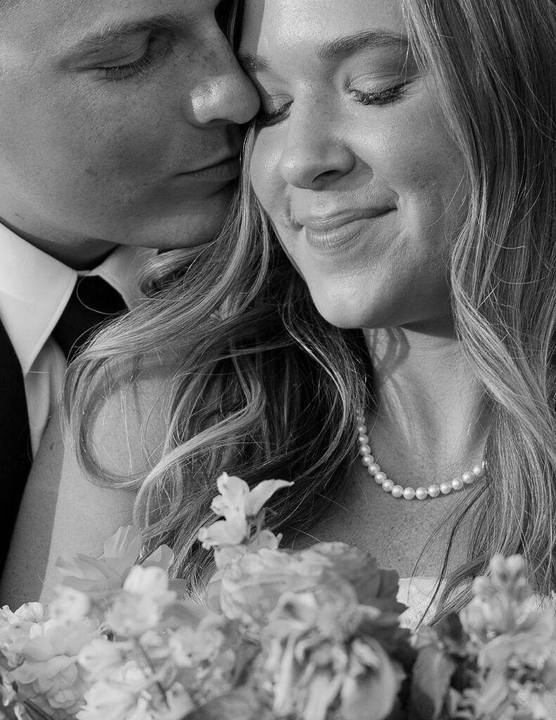 Black and white close-up portrait of bride smiling while groom kisses her cheek during an intimate wedding moment