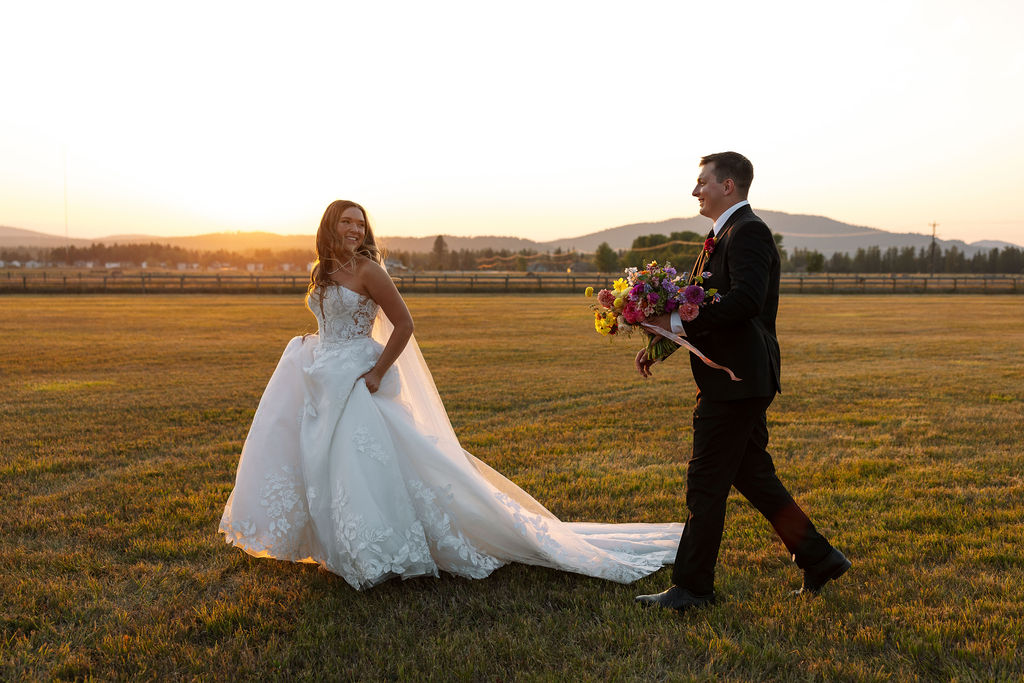 Bride and groom walking together across an open field with colorful bouquet and mountain views during a summer wedding in Whitefish Montana