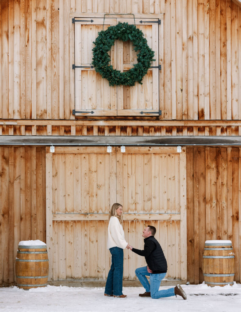 Couple during surprise proposal outside rustic barn in snowy Whitefish Montana