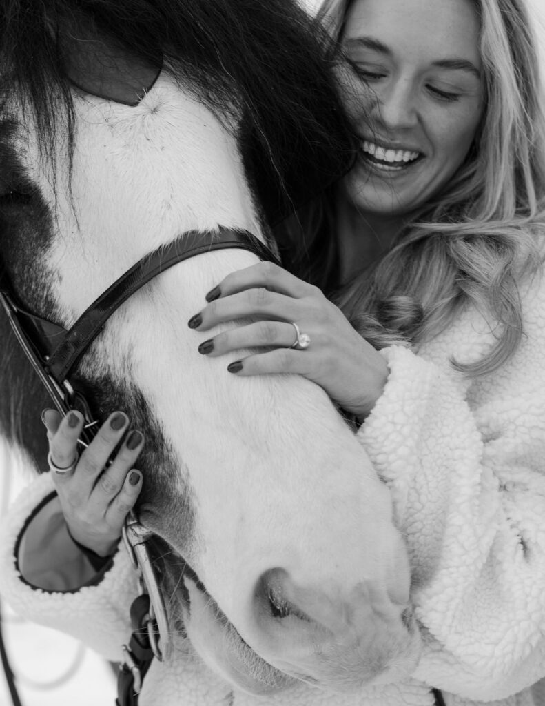 Black and white photo of couple hugging beside horse during winter engagement session in Whitefish Montana